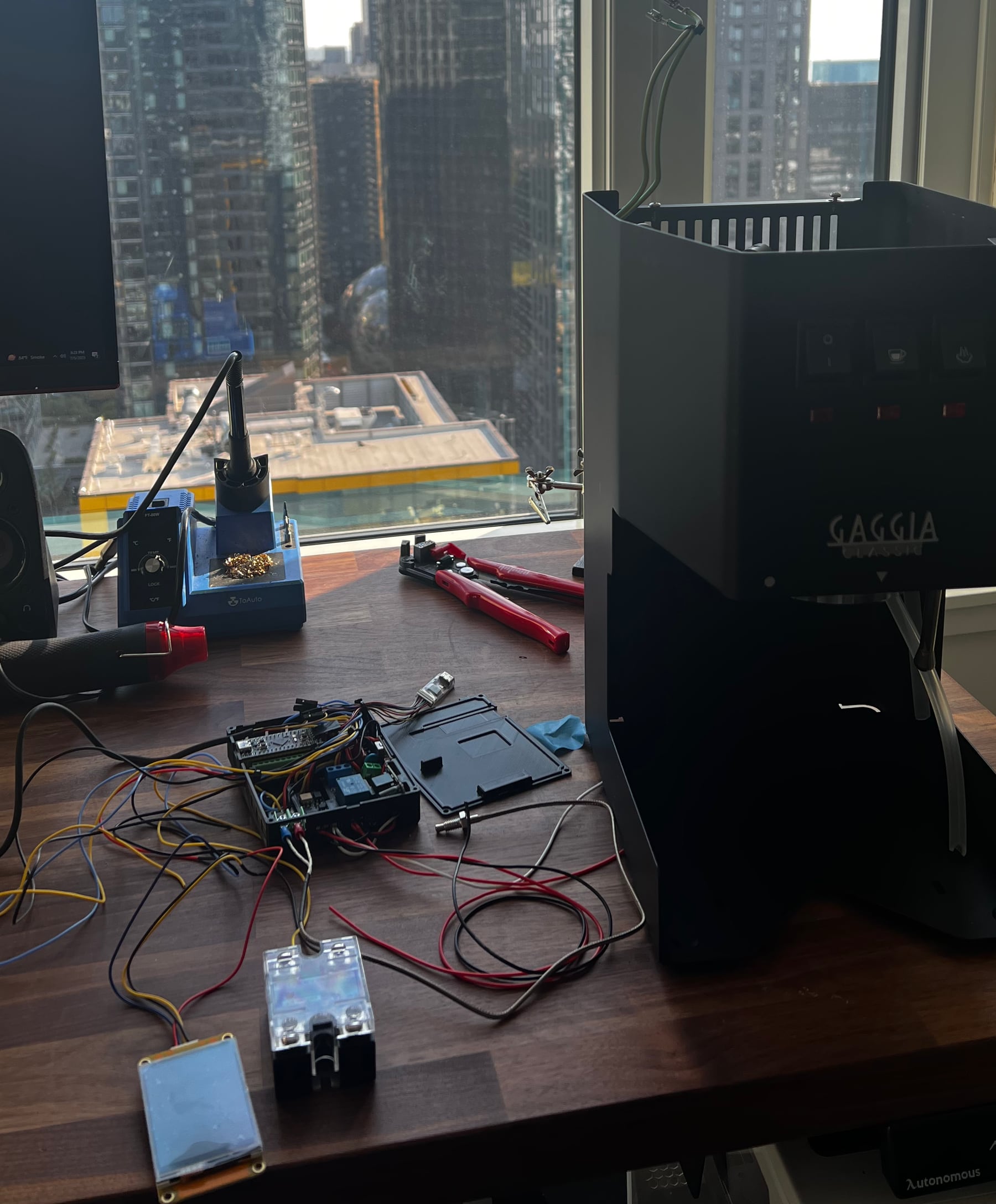 The Gaggia's shell open on a desk, surrounded by loose wiring, the relay module, and a soldering iron, mid-build, with the city out the window behind it.