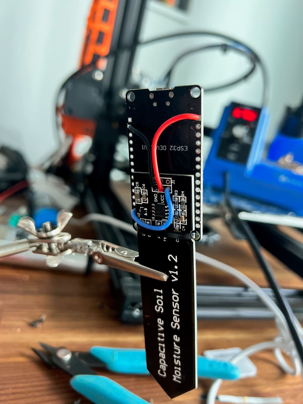 An ESP32 dev board wired to a capacitive soil moisture sensor, clipped in a helping-hands stand on a workbench.